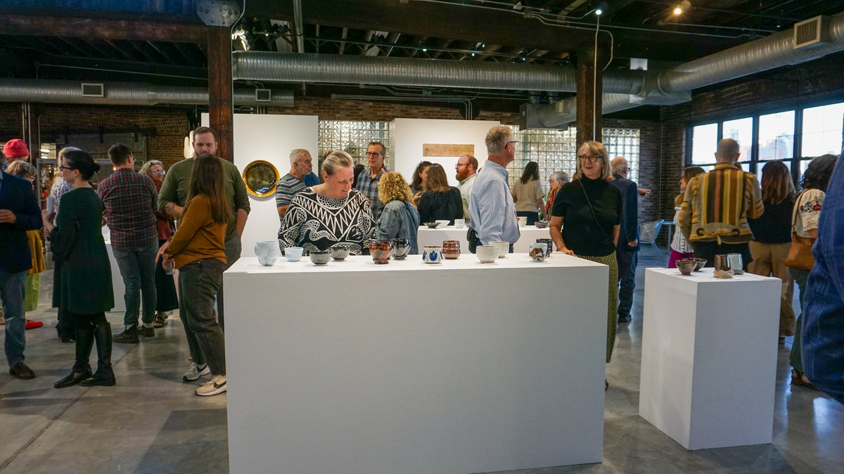 A group of people gathered around white display pedestals in a loft-style gallery or event space, viewing small tabletop objects. The scene is indoors with exposed ceiling ducts, brick walls, and large windows in the background. Victor Babu
